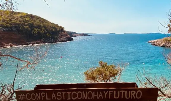 Banco de madera con el cartel #ConPlasticoNoHayFuturo frente a una cala de aguas turquesas